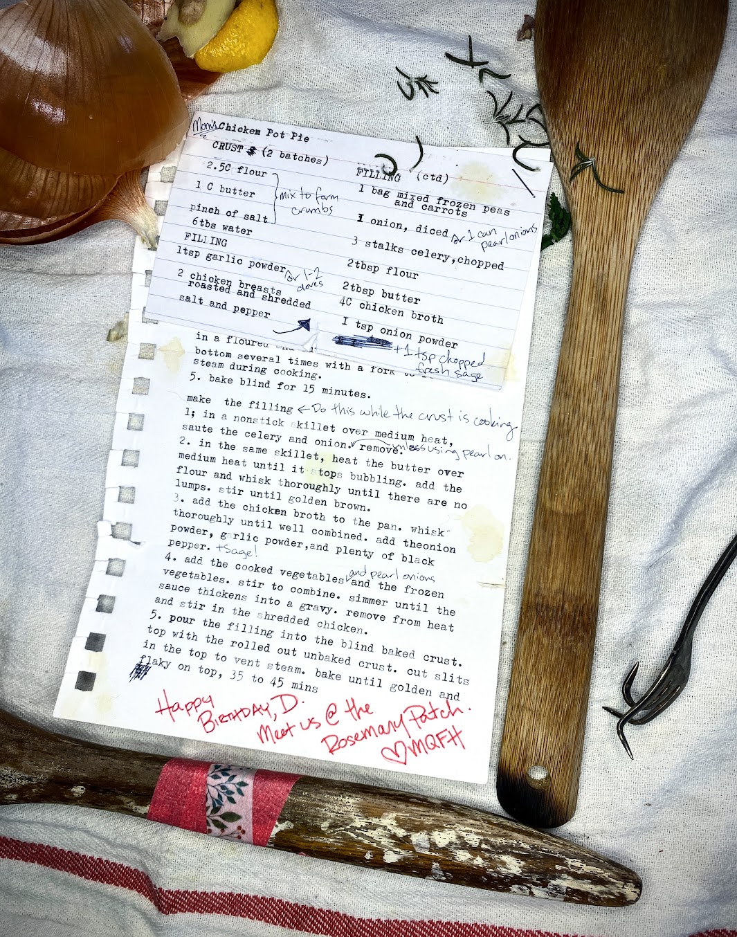 A recipe card, typewritten on an index card, stapled to a torn sheet of notebook paper with a typewritten recipe on it. Both are weathered, torn, stained, and annotated. The card is on top of a stained white kitchen towel, next to a couple of burned and repaired wooden cooking spoons, some onion and lemon scraps, a scattering of rosemary, and a bent fork. Visible recipe text is as follows (all is typewritten unless otherwise indicated; see story text for recipe in full):Index card: Chicken Pot Pie (handwritten annotation reading “Mom’s”)Crust (2 batches)2½ cups flour Pinch of salt 1 cup butter (handwritten annotation indicates these three ingredients should be mixed to form crumbs)6 tablespoons waterFilling1 teaspoon garlic powder – handwritten annotation reading “or 1-2 cloves”2 chicken breasts roasted and shreddedSalt and pepper – hand-drawn arrow points up to the next column Filling continued1 bag mixed frozen peas and carrots1 onion, diced; handwritten annotation reading “or 1 can pearl onions”3 stalks celery, chopped2 tablespoons flour2 tablespoons butter4 cups chicken broth1 teaspoon onion powder(Several typing errors are scribbled out; handwritten annotation reading “1 tsp chopped fresh sage”)The index card overlaps the recipe page. The recipe visible on the page is as follows:5. Bake blind for 15 minutes. Make the Filling (handwritten annotation indicates to do this while the crust is cooking.)1. In a nonstick skillet over medium heat, sauté the celery and onion. Handwritten note says “unless using pearl on.” Remove.2. In the same skillet, heat the butter over medium heat until it stops bubbling. Add the flour and whisk thoroughly until there are no lumps. Stir until golden brown.3. Add the chicken broth to the pan. Whisk thoroughly until well-combined. Add the onion powder, garlic powder, and plenty of black pepper. (Handwritten annotation reads “+ sage!”)4. Add the cooked vegetables (handwritten annotation reads “^or 1 can pearl onions”) and the frozen vegetables. Stir to combine. Simmer until the sauce thickens into a gravy. Remove from the heat and stir in the shredded chicken.
7. Pour the filling into the blind-baked crust. Top with the rolled-out unbaked crust. Cut slits in the top to vent steam. Bake until golden and flaky on top, 35–45 minutes.
Below the recipe, a typing error is scribbled out. A handwritten note in different handwriting from the recipe annotations, in red marker, reads: “Happy birthday, D. Meet us @ the Rosemary Patch. Heart, MQFH”